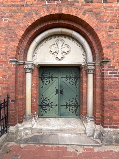 This is a close-up, eye-level, top-down shot of an ornate, historic doorway, likely part of a church or similar substantial building, located in Bassum, Germany. The primary materials visible are red brick and a light-colored, carved stone. The doorway itself is composed of two green wooden doors, featuring intricate black wrought-iron decorative elements and hinges. Above the doors, a large, round stone carving with a cross motif is set within a lighter stone surround, which is itself framed by a series of concentric brick arches. The brickwork shows some signs of age and weathering. The stone elements display detailed carvings and appear to be of a classical or gothic architectural style. The scene is well-lit, suggesting it was captured during the daytime, with no obvious indicators of specific weather conditions. The focus is entirely on the architectural details and the craftsmanship of the entrance, creating a sense of historical significance and artistry. There is no text visible in the image.