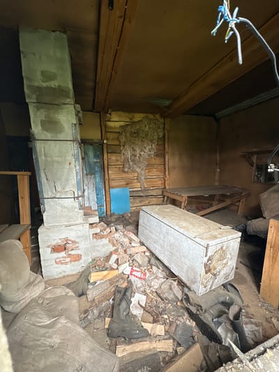This image depicts the interior of a dilapidated wooden structure, likely a shed or abandoned dwelling, in Abzakovo, Russia. The scene is cluttered with debris, suggesting neglect and disuse. A prominent feature is a tall, crumbling concrete chimney or support column on the left, partially constructed of brick. Scattered around the base of this structure are broken bricks and other rubble. In the center of the room, a large, light-colored rectangular object, possibly a chest freezer or a large storage container, sits on the floor amidst more debris. To the right, a simple wooden table or bench is visible. The walls are made of rough-hewn logs, and the ceiling is constructed of wooden planks. Various items of clothing, including boots, are strewn across the floor, along with other unidentifiable objects and trash. Hanging from the ceiling are wires and what appears to be insulation or nesting material. The overall impression is one of disarray and abandonment within a rustic, possibly rural setting.