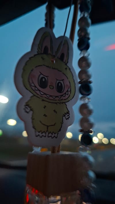 The image shows a fuzzy, monster-like sticker hanging from a beaded string. The sticker is predominantly green and cream-colored, with cartoonish features and large eyes. The beaded string consists of pearl-like white beads interspersed with dark blue beads. The background is blurred, with hints of city lights and a dusky blue sky. The photo was taken in a car. The sticker is hung in a car. It is difficult to provide an exact location within Самахвалавіцкі сельскі Савет, Belarus, as the image does not contain any distinctive landmarks or visual cues specific to a location within the village.