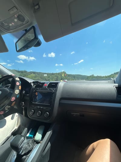 The image shows the interior of a car, looking out through the windshield. The view from the car reveals a sunny day with a blue sky and scattered clouds, overlooking a landscape of rolling hills covered in green trees. The road ahead appears to be a highway with other vehicles visible in the distance. The interior of the car includes the dashboard, steering wheel, center console with a gear shifter, and a person's legs. The steering wheel is decorated with floral patterns. The dashboard has a navigation screen, air vents, and the word "AIRBAG" is visible. In the center console, there are items like a pack of gum and what appears to be a small device. The image seems to be taken while the car is in motion. The location is identified as the city of Rajana, Bulgaria.