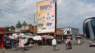 The scene is a street corner in Keshero, Democratic Republic of the Congo, located at coordinates -1.648 latitude, 29.187 longitude. There is a large billboard with a bright pink and blue advertisement for a brand of soap called “Revital” in the background. In the foreground, a man in a white shirt and khaki pants is walking towards the camera. A few other pedestrians are visible, and a man in a black jacket and a motorcycle helmet is riding a motorcycle. There are several shops and stalls in the background, including one with a striped awning. The street is paved with dirt and there are many power lines strung above. The sky is cloudy and there is a blue car in the right corner of the frame. The scene appears to be from a moving vehicle.