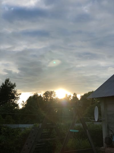 This media file captures a rural outdoor scene at sunset in Staroe Istomino, Russia. The setting appears to be a backyard or property with various structures and natural elements. Dominating the frame is a dramatic sky filled with a mix of clouds, with the setting sun breaking through, casting a warm, golden light. This illumination creates a lens flare effect in the sky and highlights the edges of the clouds.

Visible man-made objects include a section of a corrugated metal roof on the left, suggesting a building or shed. To the right, there is a wooden structure, possibly a deck or a swing set, with a satellite dish mounted on a pole. Another object, appearing to be a green hoop, hangs from the wooden structure. Dense green foliage from trees and bushes surrounds the area, some of which are silhouetted against the bright sky.

The time of day is clearly sunset, indicated by the low angle of the sun and the warm, diffused light. The weather appears to be partly cloudy. The overall mood is serene and peaceful, with the natural beauty of the sunset contrasting with the rustic, functional elements of the property. There is no visible text in the image.