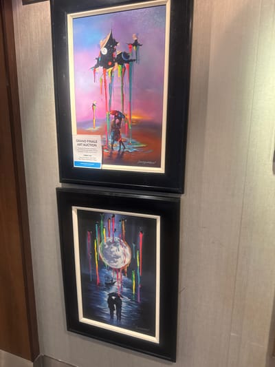 Two framed paintings are displayed on a textured, light-colored wall. The top painting features a surreal landscape with a dark, angular structure from which brightly colored paint drips. A silhouetted couple holding an umbrella is visible in the foreground. A white informational card is attached to the matting of this painting, indicating a "GRAND FINALE ART AUCTION" for a piece titled "GIBRALTAR." The bottom painting depicts a dark, watery scene with a large, luminous moon or orb in the sky. Streaks of vibrant color descend from the orb, similar to the drips in the upper painting. A silhouetted couple holding umbrellas stands on a reflective surface, and a sailing ship is visible in the distance. There are no discernible elements in the image that would allow for pinpointing a location in the city of Motril, Spain.