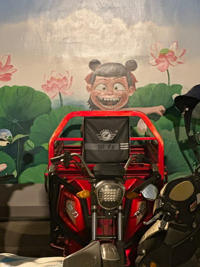 A red electric tricycle with a black cargo bag on the back is parked in front of a mural painted on a wall. The mural depicts a cartoon character with large eyes and a wide grin, peeking from behind large green lotus leaves and pink lotus flowers. The character has dark hair styled in two buns. The tricycle has a silver headlight and the cargo bag has a logo with Chinese characters. To the right of the red tricycle, a portion of a black and cream colored scooter is visible. The scene appears to be outdoors, possibly in a residential or commercial area of Eshan Yi Autonomous County, China, given the style of the mural and the presence of the vehicles.