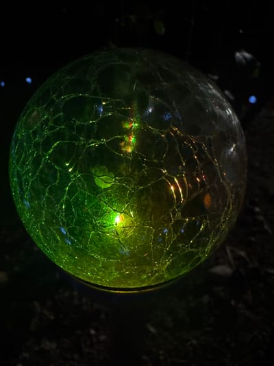 A glass orb, illuminated by internal lights, rests on a dark surface, possibly soil or foliage. The orb displays a cracked or textured surface, with green and yellow light emanating from within and reflecting off the intricate patterns. The background is dark and out of focus, with a few blurred blue lights visible in the upper left corner. The scene appears to be outdoors, possibly in a garden or yard. The specific location in Belashtitsa, Bulgaria, cannot be determined from the image alone.