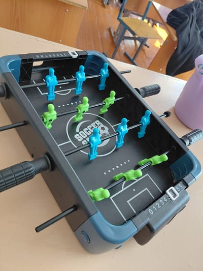 A close-up, eye-level shot captures a blue and gray foosball table set on a light brown wooden surface. The table features two rows of blue foosball players and two rows of green foosball players. A white "SOCCER GAME" logo is visible on the black playing surface, which is marked with white lines simulating a soccer field. A scoring mechanism with numbers 0 through 9 is attached to the right side of the table. In the background, a blurry classroom setting is visible, with wooden desks and chairs. The scene is devoid of people, and the overall lighting suggests an indoor environment. The image does not contain any information to place it in Svetlogorsk, Russia.