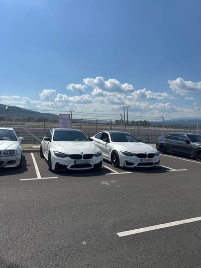 The image captures a sun-drenched outdoor parking lot, likely at the race track in Dospey, Bulgaria, given the background. Four BMW cars are parked in designated spots. The cars visible are a silver BMW on the far left, followed by two white BMWs, and a dark gray BMW on the far right. The white BMWs are the most prominent, with one displaying a license plate that reads "CB 6566 MO" and the other "CO 8881 BX". The ground is paved with asphalt and marked with white parking lines. In the background, a metal fence separates the parking area from a race track, with hills and a blue sky with scattered white clouds completing the scene. There are no people visible in the image.