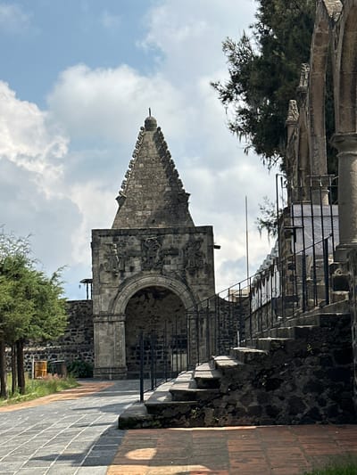 The image shows a stone structure with an arched entrance and a pointed spire, located in San Andrés Calpan, Mexico. The architecture appears to be historic, with intricate carvings above the archway and decorative elements along the spire's edges. To the right of the main structure, a stone staircase with a wrought-iron railing ascends. The foreground features a paved pathway composed of large, square tiles in varying shades of gray, bordered by a reddish-orange tile pathway. Lush green trees frame the scene, and the sky above is a mix of blue with scattered white clouds. The environment appears to be an outdoor historical site or landmark.