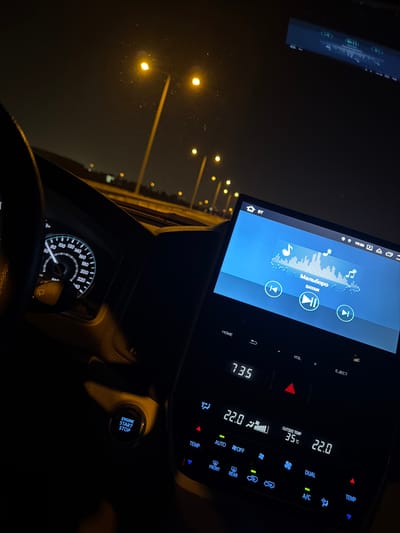 The image captures the interior of a car at night, facing forward from the driver's perspective. The scene is dark, illuminated primarily by the car's interior displays and distant streetlights visible through the windshield.

The primary subjects are the car's infotainment system, which dominates the right side of the frame, and a portion of the steering wheel and speedometer on the left. Outside, a road or highway with bright, warm-toned streetlights stretches into the distance, suggesting the car is stopped or driving slowly in an urban or suburban area.

On the large infotainment screen, music is playing, with Cyrillic text "Мальборо" (Malboro) and "SAYAN" visible as the artist/song title. A clock on the screen shows "19:36". The climate control section of the display shows an "OUTSIDE TEMP 35°C" and cabin temperature settings of "22.0°C" for both sides, indicating a dual-zone system. Other climate controls like "A/C," "AUTO," "DUAL," and fan speed icons are also visible. A digital time "735" is shown on the climate control panel.

Part of the speedometer is visible on the dashboard, with the needle resting at 0 km/h, suggesting the car is stationary. Below the steering wheel, an "ENGINE START STOP" button is clearly lit. The overall setting appears to be a nighttime drive or stop in Umm Salal Ali, Qatar, as indicated by the high outside temperature.