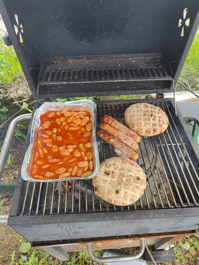 An overhead shot captures a black, well-used barbecue grill actively cooking food outdoors in Bad Pyrmont, Germany. The grill, with its lid open, reveals a variety of items being prepared over visible embers.
On the left side of the grill grate, a rectangular aluminum foil pan holds a generous portion of baked beans in a rich, orange-red tomato sauce. The beans appear to be simmering, indicating they are being heated or kept warm. To the right of the beans, several hot dogs, each wrapped in strips of bacon, are grilling. The bacon shows signs of cooking, with some browning and crisping. Two round, flatbread-like items, possibly naan or pita bread, with distinct grill marks and a lightly browned, speckled surface, are placed on the grate – one near the upper right and another towards the lower center.
The grill itself is made of dark metal, likely black, showing signs of regular use with some residue on the interior surfaces and grate. Decorative cutouts resembling a stylized cross or propeller shape are visible on the interior sides of the grill's open lid. The lower structure of the grill appears somewhat rustic, with traces of rust on its metal legs and a wheel visible on the right side, suggesting it is portable.
The setting is outdoors, likely a garden or backyard, with green foliage, grass, and a chain-link fence visible in the background, particularly on the left side. The ground beneath and around the grill is unpaved earth with scattered weeds. A silver can with illegible text is partially visible on the ground below the grill's handle on the left. The scene conveys a casual, daytime outdoor cooking event.