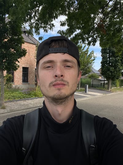 A medium shot selfie is taken outdoors in Diepenbeek, Belgium, on a paved road with a tree-lined background. The subject is a young Caucasian male with light skin, brown eyes, and short brown hair, wearing a black baseball cap and a black turtleneck shirt. He has a light beard and mustache and a few freckles on his face. He is looking directly at the camera with a neutral expression. He is also wearing a black backpack. In the background, there is a red brick house with black-framed windows, a garden with green plants and purple flowers, and several trees. Further in the distance, there are more houses with solar panels on their roofs and a trash bin on the side of the road. The sky is blue with some white clouds.