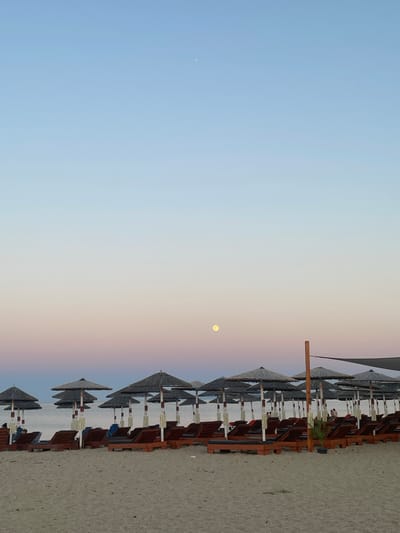 This is a photograph of a beach in Nea Potidea, Greece. The image depicts a row of empty lounge chairs and thatched umbrellas set up on a sandy beach. In the background, the calm sea is visible, with a gentle gradient of colors in the sky transitioning from a pale blue at the top to soft pinks and oranges near the horizon. The moon is visible in the sky. The overall scene suggests a peaceful and serene atmosphere, likely taken during sunrise or sunset.
