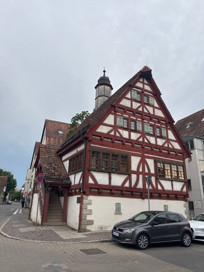 The image depicts a street scene in Sindelfingen, Germany, focusing on a prominent half-timbered building with a red and white facade. The building features multiple windows with wooden frames and green shutters on its upper levels. A steep, external staircase with wooden supports leads to an entrance on the left side of the building.
In the foreground, a dark gray Volkswagen Polo is parked on the street, facing left. To the right of the Polo, a portion of a white car is visible. The street is paved with cobblestones and bordered by sidewalks. A circular sign with a red border and a blue arrow pointing left, indicating a no-entry zone or a specific traffic direction, is affixed to the building above the staircase. A "30" speed limit sign is also visible on the left side of the street.
The sky is overcast and gray. In the background, other buildings and trees can be seen along the street. The overall impression is of a historical European town.