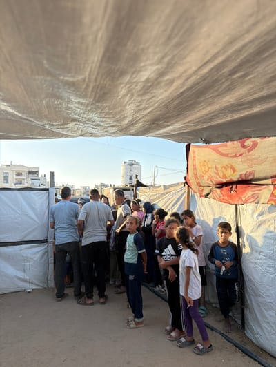 A group of people, including men, women, and children, are gathered outdoors in what appears to be a temporary shelter in Jabalia, Palestinian Territories. The shelter is covered by a large, light-colored tarp, and a patterned textile is hanging from the side. In the background, buildings and a water tank are visible, suggesting an urban or semi-urban environment. Some of the people are standing and looking around, while others seem to be in a line, possibly waiting for something. The ground is sandy, and the lighting suggests it is daytime, possibly late afternoon due to the warm light.