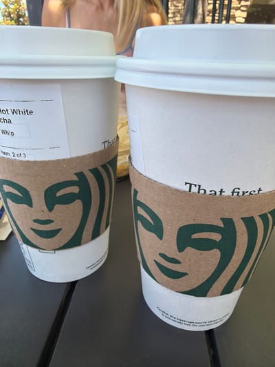 Two Starbucks coffee cups are placed on a dark gray table. The cup on the left has a white label with black text that reads "Hot White Mocha Whip" and "Item: 2 of 3". Both cups have brown cardboard sleeves with the Starbucks siren logo printed in green. The cup on the right has text that reads "That first". The background shows a person with blonde hair and a striped top, and a stone wall behind them. The scene is outdoors.