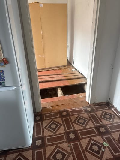 The image shows an interior view of what appears to be a room undergoing renovation or in disrepair. In the foreground, a portion of a checkered linoleum floor with brown and tan geometric patterns is visible. To the left, a white refrigerator is partially in frame. An open doorway leads to a room where the wooden floorboards have been partially removed, revealing a dark cavity beneath. The exposed wooden joists and floorboards are weathered and show signs of age and use, with some planks appearing reddish-brown and others a lighter wood tone. In the background, a large, light brown cabinet or wardrobe stands against a wall. The walls in the scene are painted white and show some cracking and discoloration. The overall environment suggests an indoor domestic setting, possibly a kitchen or hallway, with a clear indication of structural work or damage to the floor. There are no people or animals visible in the image. The location is not specified as being in Yabogan, Russia.