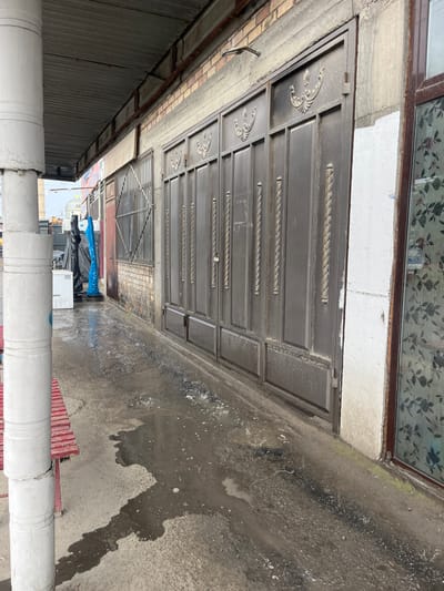 This is an outdoor, ground-level, medium shot of a wet pathway leading to a large, dark metal gate. The gate is composed of several panels, each with a decorative top section and vertical rope-like designs. To the left of the gate is a section of brick wall and a smaller metal gate with vertical bars. Further left, a white pillar stands in the foreground. Beyond the pillar, a red bench is visible. In the background, there are two figures, one in black and one in blue plastic wrapping, standing near a white appliance. The ground is covered in puddles of water, indicating recent rain or a water spill. The environment appears to be a commercial or industrial area in Medrese, Kyrgyzstan, with a covered walkway and a building facade. The lighting is overcast, suggesting a cloudy day.