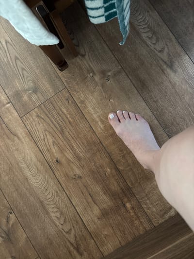 A close-up, high-angle shot shows a person's bare foot with white-painted toenails resting on a wooden floor. The floorboards are light brown with a visible wood grain pattern and appear to be laminate. To the left of the foot, a corner of a blue and white checkered blanket is visible, draped over the edge of what appears to be a wooden chair or table leg. Further to the left and below the blanket, a white fuzzy material is also present, possibly from a rug or another blanket. The lighting suggests an indoor setting, with no specific time of day or weather information discernible. The image is focused on the foot and its immediate surroundings, creating an intimate and casual feel. There is no visible text in the image.