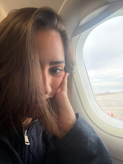 A woman is seated in an airplane by the window. The view outside the window suggests the plane is on the ground or taxiing, with possible airport infrastructure visible. The woman is resting her face on her hand, looking towards the camera. She is wearing a dark jacket or sweater with a visible zipper pull, potentially branded with "POLO". The overall environment appears to be inside an airplane cabin. There is no information to specify its location in San Antonio.
