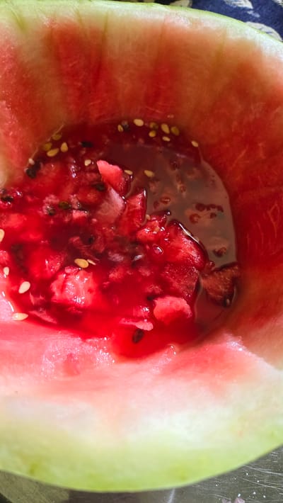 The image shows a close-up of the interior of a cut watermelon. The rind, a pale green fading to white, forms the outer edge of the frame. The fleshy interior is bright red, with some white pith visible near the rind. Small, dark seeds are scattered throughout the red flesh, and some have sunk to the bottom where juice has collected, creating a pool of deep red liquid mixed with finely diced watermelon pieces. There are no people, buildings, or any indication of a specific location within Mary City, Turkmenistan. The scene is solely focused on the fruit itself, presented in a close-up, almost abstract manner.