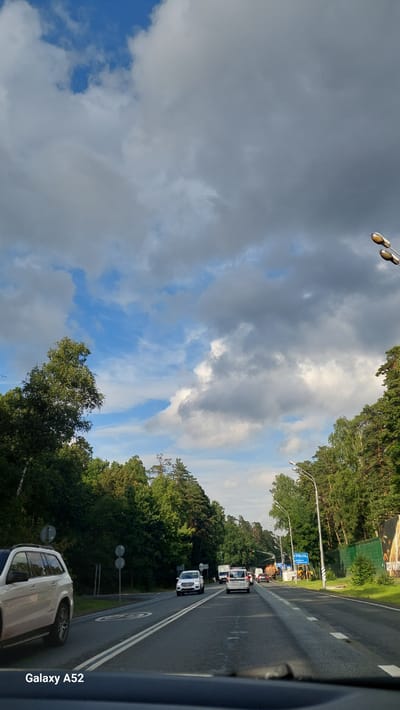 The image shows a view from a vehicle driving on a road lined with trees on both sides. The sky is partly cloudy with patches of blue sky visible. Several cars are on the road, including a white SUV in the foreground on the left and a white car further ahead. There are also streetlights and traffic signals visible. The road appears to be a multi-lane highway. The text "Galaxy A52" is visible in the bottom left corner of the image, indicating the device used to capture the photo. The scene appears to be in a suburban or rural area, possibly on the outskirts of a city. The overall impression is of a typical summer day drive.