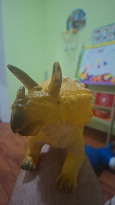 A yellow toy triceratops dinosaur is positioned in the foreground of the image, its head facing left. The dinosaur is resting on a surface that appears to be a person's leg. In the background, a green wall is visible, with a blurred basketball hoop and backboard hanging on it. To the right of the hoop, a colorful whiteboard or easel with magnetic letters is also visible. The floor in the background is made of wooden planks. The overall scene suggests an indoor setting, possibly a child's room or playroom. The image does not provide any information about the city of Voinova, Moldova.