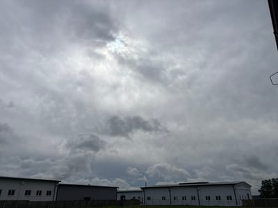 The image shows a view of several modern buildings under a cloudy sky. The buildings appear to be low-rise industrial or commercial structures with white facades and dark roofs. They are arranged in a row, with some spaces between them. The sky is predominantly overcast with thick, gray clouds, but there are areas where the sun is breaking through, creating a bright, diffused light effect. A dark metal structure, possibly part of a fence or railing, is visible on the far right side of the frame, extending downwards. In the foreground, a strip of green grass is visible in front of the buildings. The overall impression is one of an industrial or commercial area on an overcast day.
