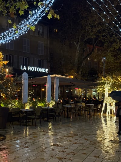 A nighttime view of the restaurant "La Rotonde." Tables and chairs line the outdoor patio, some covered with white cloths. The pavement is wet, reflecting the light from strings of lights and illuminated trees. The restaurant sign glows brightly against the building facade. Decorative light displays add to the festive atmosphere. A person holding an umbrella is visible near the top.