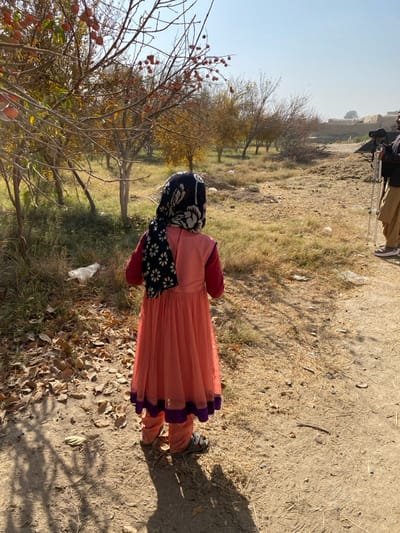 The image depicts a rural scene in Bazar-e Panjva'i, Afghanistan, under clear, bright daylight. A young child, viewed from the back, stands on a dirt path, facing forward towards an orchard. The child wears a long-sleeved salmon-colored dress with a purple trim at the hem, over what appears to be matching trousers, and open-toed sandals. A black headscarf with white floral patterns covers the child's head and upper back.

To the right and slightly behind the child, an adult male is partially visible, wearing light-colored traditional clothing (trousers) and operating a camera mounted on a tripod.

The immediate foreground consists of a dirt path with scattered brown leaves. To the left, a small orchard features deciduous trees with sparse reddish-brown foliage, some bearing visible red fruits, consistent with autumn. The ground beneath the trees is covered in dry grass and fallen leaves, with some light debris including a white plastic bag. The background reveals more trees and indistinct, light-colored structures, possibly mud-brick buildings, under a pale blue, clear sky. Strong shadows are cast on the ground, indicating direct sunlight.