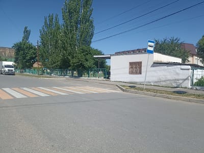 A low-angle, eye-level shot captures a street scene in Izberbash, Russia. In the foreground, a grey asphalt road features a pedestrian crossing with alternating orange and white stripes. To the left, a white truck with a grey trim is partially visible, parked on the left side of the road. A green metal fence runs along the background, separating the road from a row of tall green trees and a low, white building with a brown-tiled roof. A blue and white bus stop sign is mounted on a pole to the right of the building. The sky is a clear, bright blue.