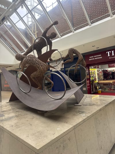 The image depicts an indoor scene within a commercial shopping center in Woking, United Kingdom, centered around a large public art sculpture. The sculpture is composed of three stylized human figures made from interwoven dark brown natural fibers, each in a dynamic, athletic pose. These figures interact with multiple circular silver-colored hoops and a large, dark bronze-colored, undulating metal form that serves as the sculpture's primary structure. The entire artwork is situated on an elevated plinth of light beige, mottled stone tiles.
An overhead glass skylight, framed in white, reveals an external reddish-brown brick building with metal structures, including what appears to be a fire escape, visible against an overcast sky. Retail units line the periphery of the space. To the right, a Timpson store with a red facade and white lettering displays 'SHOE REPAIRS WATCH REPAIRS' above its brightly lit interior, which features various retail products. To the left, a retail unit secured by a blue roller shutter shows signage for 'Charity Bookshop The Trus...', while an adjacent unit is secured by a black roller shutter. The floor throughout the area is surfaced with matching light beige stone tiles, exhibiting subtle reflections. The space is illuminated by a combination of natural light from the skylight and artificial ceiling lighting.