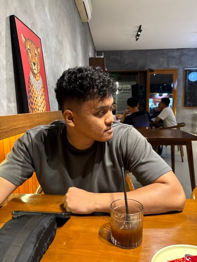 A man with curly dark hair sits at a wooden table with a glass of iced coffee. He wears a gray t-shirt. A black bag rests on the table. A framed picture of a leopard in pearls is on the wall nearby. In the background, other people sit at tables in a cafe setting. There is a white wall and what looks like an air conditioner. The image is slightly tilted.