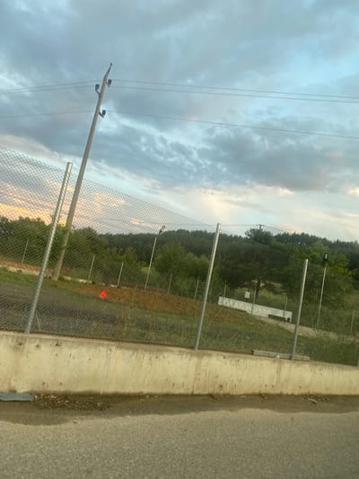 The image depicts an outdoor scene, likely in the vicinity of Ablanitsa, Bulgaria, under a cloudy sky. In the foreground, a paved surface, possibly a road or pathway, is visible, bordered by a concrete barrier. Beyond the barrier, a chain-link fence runs horizontally across the frame, separating it from the background. The fence is supported by metal posts.
Behind the fence, the terrain slopes upwards, covered in grass and dirt. Scattered throughout this area are various objects including what appear to be construction materials or debris, and some red markers or flags. A cluster of trees and dense foliage forms a natural backdrop, with a more distant line of trees suggesting a forest or heavily wooded area. Two prominent utility poles, one leaning significantly, are visible against the sky, with wires extending between them. One of the utility poles has what appear to be insulators attached. Further in the distance, a white structure or building can be discerned amidst the trees, and a light fixture is mounted on a pole. The overall lighting suggests it is either early morning or late evening, given the soft illumination and the color of the sky. There are no people or animals clearly visible in the image.