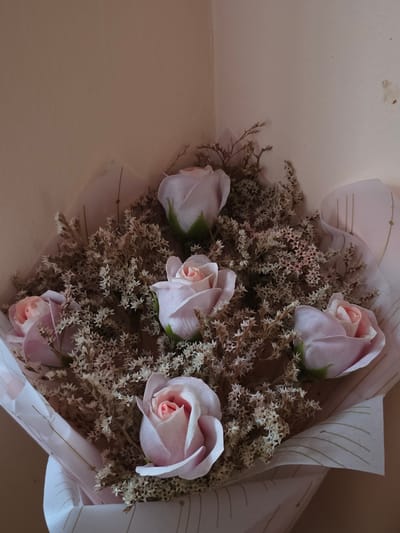 This medium close-up image, taken in Kalitinovo, Bulgaria, features a bouquet composed of five artificial or dried roses and a substantial amount of dried, wispy filler flowers. The roses are a soft pastel pink or light purple color, with some showing a deeper pink at their core. They are nestled among a dense arrangement of smaller, beige-toned dried floral material, possibly statice, which has a delicate, star-like appearance.
The bouquet is encased in light-colored wrapping paper, likely off-white or pale pink, adorned with subtle, faint, and irregular vertical striped patterns. The paper is loosely folded around the flowers, creating a natural, gentle presentation. The entire floral arrangement rests on a smooth, uniformly light-beige surface, such as a table or floor, and is positioned against a plain wall of a similar light hue. A small, indistinct dark mark is visible on the upper left portion of the wall.
The lighting is soft and diffused, characteristic of an indoor setting with ambient illumination, casting gentle shadows that highlight the textures of the flowers and wrapping. There is no specific activity taking place, as the image is a still life of the bouquet. The overall impression is one of calm and delicate beauty. No legible text is visible within the frame, only the decorative linear patterns on the wrapping paper.