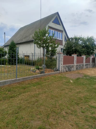 A two-story house with a grey asphalt shingle roof is visible, situated in Nosivka, Ukraine. The house has light-colored brick walls and features several windows, including two triangular windows on the upper gable. A metal fence with a concrete base runs across the foreground, with a second, more decorative fence constructed from stone and wood panels further back. Several trees and green bushes are present in the yard, along with a small patch of dry grass. The sky is overcast with clouds.