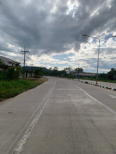 The image depicts a two-lane concrete road in the Mae Chan District of Thailand, likely during a cloudy day with sunlight breaking through the overcast sky. The road appears to be newly constructed or recently resurfaced, with distinct lane markings. To the left of the road, there is a grassy verge with lush green vegetation. Beyond the verge, a building with a white facade and a dark roof is visible, along with utility poles and overhead wires. Further in the background, more buildings with various roof types and trees line the sides of the road, leading towards a forested mountain range under a dramatic sky filled with dark and light grey clouds. Streetlights are positioned along the right side of the road, which has a black and white striped barrier marking its edge. The overall scene suggests a semi-rural or suburban setting within the Mae Chan District.
