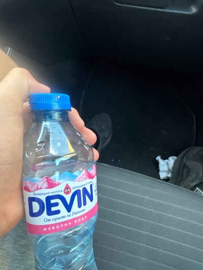 A hand holds a partially empty water bottle labeled "Devin" in Bulgarian, with a blue cap, inside a car. The car's interior consists of a dark dashboard, black floor mats, a gray striped seat, and a shoe on the floor. There is no information to support a location in Kaloyanovo, Bulgaria.