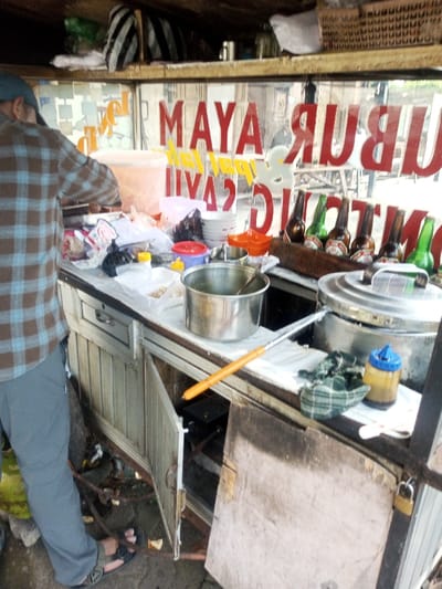The image depicts a street food stall, observed from a slightly elevated perspective, with a person standing behind the counter. The stall features an aluminum-framed structure with a stainless steel countertop. A lone individual, seen from the back, wears a blue and brown plaid long-sleeved shirt, grey pants, and dark sandals, actively engaged in preparing or serving food on the left side of the counter.

The counter is cluttered with various items. On the left, a large, translucent plastic container holds an orange substance. Numerous white plastic bowls are stacked, alongside smaller orange and red plastic containers. A large open metal pot with a ladle rests centrally, next to a second, lidded metal pot. A long-handled utensil with an orange grip extends from the right side of the counter. Towards the right, several brown and green glass bottles, resembling beer bottles, are arranged. A small condiment bottle with a blue cap and a folded green plaid cloth are also present. The ground beneath the stall is concrete, and a green gas cylinder is visible near the person's feet. An open cabinet door on the stall's lower section reveals a gas burner and associated tubing. A weathered wooden panel is secured to the right side of the stall with a padlock.

Through the clear glass partition above the counter, mirrored red text spelling "BUBUR AYAM" and yellow text reading "pedas / ori" are prominently displayed, indicating "Chicken Porridge" and "spicy / original" in Indonesian. This linguistic detail suggests the scene is located in an Indonesian-speaking region, rather than Phonengam, Laos as initially indicated. The background through the glass shows a blurred urban environment.