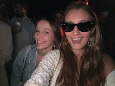 Two adult females are prominently featured in a close-up within a dimly lit social gathering in Castêlo da Maia, Portugal. The female on the left has light brown hair, is wearing a light blue denim jacket over a light-colored top, and is smiling broadly, exposing her teeth. The female on the right has long brown hair, is wearing black Wayfarer-style sunglasses, large ornate gold-colored oval hoop earrings, and a small cross pendant on a gold chain; she is also smiling with an open mouth, revealing her teeth. She wears a light-colored, patterned top, and a gold chain from an accessory is visible. Her right shoulder is bare. In the blurred background, a bearded male wearing a light-colored garment is visible over the left female's shoulder, holding a white cup. Other indistinct figures are partially visible in the low-light environment. The scene is illuminated primarily by a direct light source, highlighting the two foreground subjects against the dark background.