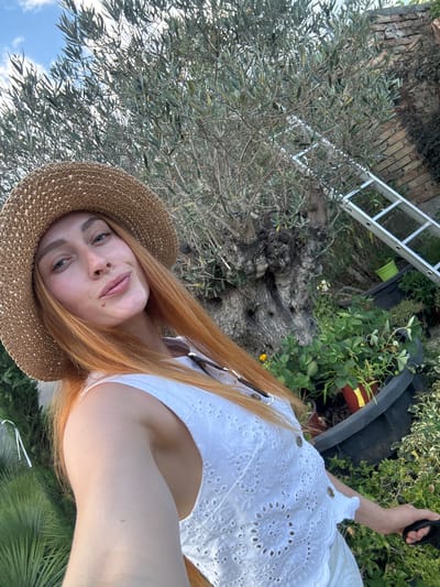 A woman with long, ginger hair and a straw hat is taking a selfie outdoors in a garden. She is wearing a white, eyelet top and is holding a black leash in her right hand. Behind her, there is a large, gnarled olive tree, a silver ladder leaning against a brick wall, and several potted plants, including strawberries. The scene appears to be located in Radanovići, Montenegro. The sky is blue with some clouds.