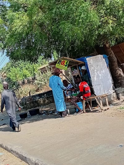 The image depicts a daytime street scene in Sharada, Nigeria. A paved road with a raised curb occupies the lower half of the frame. On the right, a makeshift vendor's stall, featuring a blue tarp roof, white side paneling, and a prominent yellow sign indicating "P.O.S Available," is positioned beneath a large, leafy green tree. Various small items are visible on shelves within the stall.
Four adult males are present. In the left midground, a man in a dark grey traditional tunic (kaftan) and cap walks away from the camera, heading left. Centered, a man in a light blue traditional tunic and cap stands with his back mostly to the camera, facing the stall. To his right, a man in a red t-shirt and dark trousers sits on a simple wooden stool, engaged with the stall. A fourth man, wearing a white cap, is partially visible behind the stall's counter, seemingly attending to customers. On the ground near the curb, two dark, round pots are visible amidst minor debris. The background shows sparse vegetation and faint structural elements under a clear sky.