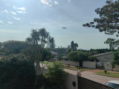 This is a daytime photograph taken from a slightly elevated vantage point, possibly a balcony or upper window, overlooking a suburban street lined with houses and lush vegetation. The sky is a pale blue with scattered white clouds, suggesting fair weather. Sunlight streams down, casting subtle shadows. In the distance, an airplane can be seen flying across the sky, heading towards the left side of the frame.
The foreground features dense green foliage, obscuring part of the view. Beyond this, a paved road curves through the neighborhood. On the right side of the road, a light-colored house with a gabled roof is visible, partially enclosed by a white brick wall and a metal gate. A car is parked behind the gate. The house has a light beige exterior and a white trim. Adjacent to the house, a smaller, light-colored structure, possibly a garage, is also visible. A small, neatly trimmed bush sits on the lawn.
On the left side of the street, an aloe tree with its characteristic thick, branching trunk and spiky leaves stands prominently. More trees and shrubs are scattered throughout the scene, including palm trees further in the background, contributing to a tropical or subtropical feel. The overall impression is one of a peaceful, well-maintained residential area on a sunny day. There is no visible text in the image.
