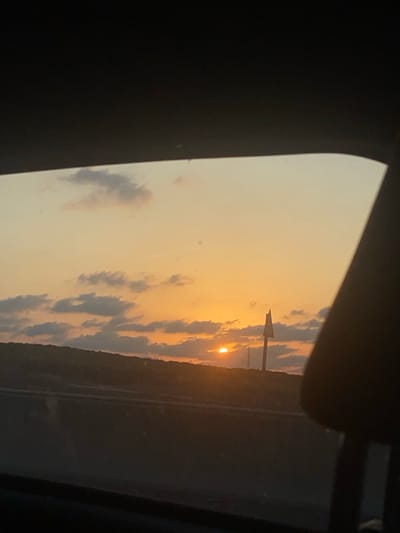 The image presents a sunset viewed from the interior of a vehicle in Beit Yizhaq - Shaar Hefer, Israel. The sky displays a gradient of orange, yellow, and pale blue, with scattered cumulus clouds illuminated by the setting sun. The sun, a distinct orange-yellow disc, is situated low on the horizon, partially obscured by both clouds and a dark, undulating landmass, likely a hill or vegetated terrain, that stretches across the mid-ground. To the right of the sun, a tall, slender pole-like structure featuring a pointed or triangular top is silhouetted against the bright sky, alongside what appears to be faint power lines or utility towers. The immediate foreground outside the vehicle shows a dark, horizontal band, suggesting a road or roadside. Inside the vehicle, the upper portion of the frame shows the dark headliner, while the right side is partially occupied by a dark, curved object consistent with a car seat's headrest. No individuals or direct actions are visible.