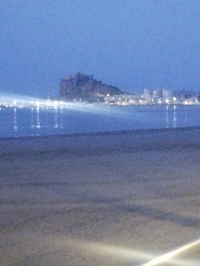 The image depicts a dusky coastal scene with a town lit up in the distance. A prominent rocky promontory, likely Isla de las Larrocas, juts out into the sea, overlooking the town and its lights. The town is characterized by buildings and illuminated structures, suggesting a populated area. The sea reflects the lights, creating a shimmering effect on the water's surface. The foreground is a dark, sandy expanse, possibly a beach, with faint streaks of light suggesting artificial illumination or reflections. The overall atmosphere is serene and tranquil, indicative of twilight or early evening.