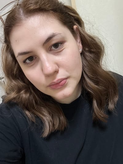A close-up, head-and-shoulders shot captures a young woman with medium-length, wavy brown hair, wearing a black t-shirt. Her right eye is slightly red, and she has a single clear stud earring in her right ear. Glasses are perched on top of her head, partially obscuring her forehead. The background is a plain, off-white wall. The lighting appears to be artificial, casting subtle shadows. There is no identifiable environmental context suggesting a specific location within Novoe Devyatkino, Russia.