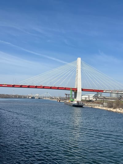 The image depicts a panoramic view of the Agigea Bridge, a cable-stayed structure spanning the Danube-Black Sea Canal in Agigea, Romania, under a clear blue sky with minimal wispy clouds. The central feature is the main bridge, characterized by a tall, slender, light-grey concrete pylon on the right. Numerous white support cables radiate from the pylon to a prominent red deck structure, which bears the white lettering "TIGITRANS." A single red vehicle is partially visible on the bridge deck.

Below the main bridge, on the right bank of the canal, a dark-hulled barge or vessel is moored. This barge is equipped with a large, bright green mobile crane positioned near its front. The canal water is a deep blue, showing rippling textures. The right shoreline consists of rocky riprap with sparse, dry vegetation. Beyond this, a series of industrial structures are visible, including a large white domed building and cylindrical silos. A rusty brown metal framework runs parallel to the canal bank.

In the mid-ground, further down the canal to the left, several other bridges are visible, including a truss bridge and another distant bridge-like structure. Adjacent to these, a lock or dam system is discernible. The distant left bank features additional industrial elements, including a tall lattice tower. No individuals are clearly discernible in the scene.