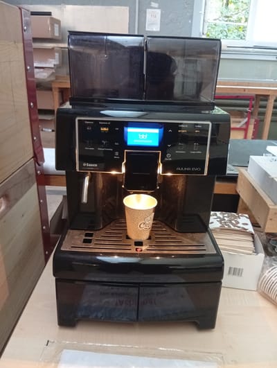 An indoor, medium shot captures a black Saeco Aulika Evo espresso machine with dual bean hoppers. The machine is positioned on a light-colored countertop. A single paper cup, featuring a patterned design and the word "Coffee," is placed on the drip tray, containing what appears to be a hot beverage. To the right of the machine, a box of disposable coffee cups and a stack of coffee coasters are visible. In the background, shelves stocked with cardboard boxes and wooden structures suggest a storage or workshop environment. The scene is illuminated by overhead lighting. No individuals are present in the image, and there is no indication of the specific location within Strengelbach, Switzerland.