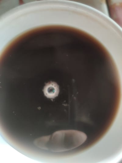 The image shows a close-up, overhead view of a dark liquid, likely coffee or tea, in a white container. The liquid fills most of the frame, with the white rim of the container visible along the top and right edges. In the center of the liquid, there is a small, iridescent bubble, reflecting light and showing hints of green and blue. There are also a few faint specks floating on the surface of the liquid. The overall lighting is soft, suggesting an indoor setting. The image does not contain any information that would allow for its localization in Ternovka, Moldova, or any other specific geographic location.