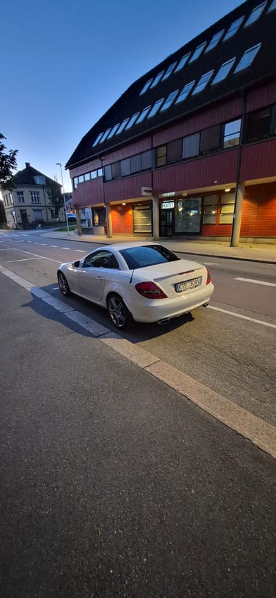 This is a vertical, medium-shot, exterior photograph captured in the city of Lillehammer, Norway. The image depicts a white Mercedes-Benz SLK convertible parked on the side of a paved road. The car has its license plate visible, which reads "KSP 308." The road has white painted lines indicating a crosswalk.

In the background, a modern building with a red facade and dark-tinted windows stands prominently. Above the windows, multiple skylights are visible on a dark, sloped roof. Signage on the building indicates "POLITI" (Police) and "HOVEDINNGANG" (Main Entrance), suggesting it is a police station. To the left of the police station, a more traditional, lighter-colored building with visible windows and a dark roof is partially visible. The sky above is clear and blue, indicating it is either early morning or late evening. The overall scene is still and quiet.
