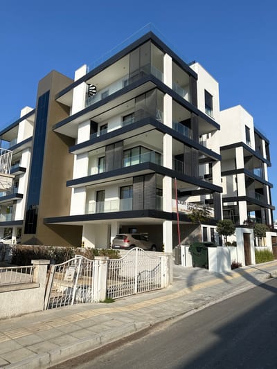The image captures a modern, multi-story building under a clear blue sky on a bright, sunny day in Egkomi Lefkosias, Cyprus. The building exhibits contemporary architecture with a facade primarily composed of stark white walls contrasted by prominent dark grey or black horizontal and vertical structural elements and balcony frames. Each level features balconies with clear glass railings, and some are equipped with dark, slatted privacy screens or louvers.

At the ground level, a covered parking area is visible, housing a silver car. An ornate white metal gate stands open, providing access to the property. Adjacent to the building, a paved sidewalk with yellow tactile paving runs parallel to a street. Green planters with small trees and shrubs are placed near the building's entrance, and a green utility bin is also present.

A sign near the building's entrance prominently displays the text "LUXURY APARTMENTS," followed by partially visible contact information including a telephone number starting with "22 22...". The scene suggests a high-end residential complex in an urban or suburban environment.
